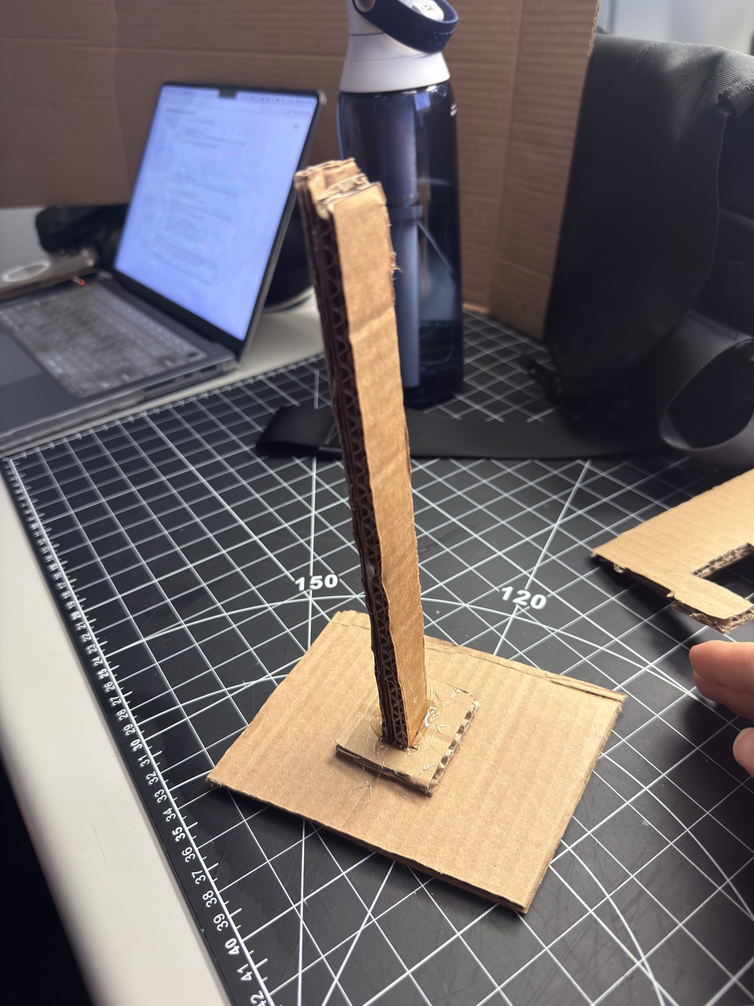 Step 1 — V1 skeleton: cardboard base + slim post to quickly test center of mass and load. Issue: wobble at the post–base joint; fix: enlarge base or add triangular bracing. Materials: corrugated cardboard, light hot-glue tacks.